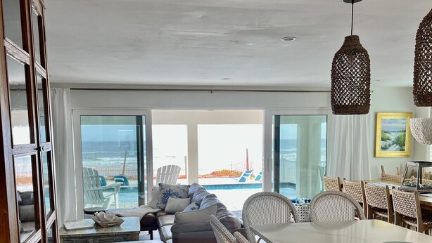 A spacious living area with a large window overlooking the ocean, a dining area with wicker furniture, and a view of the beach. - Renovated, direct oceanfront 5BR/6BA private pool home sleeps 16. (New Smyrna Beach)