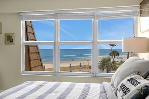 A bedroom with a large window overlooking a beach, a bed with striped bedding, and a lamp on the nightstand.