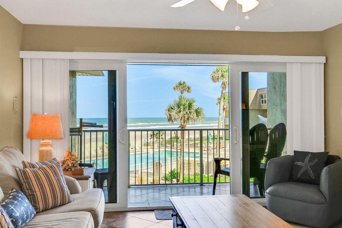 A room with a view of the beach, a balcony with a table and chairs, and a sofa with decorative pillows.