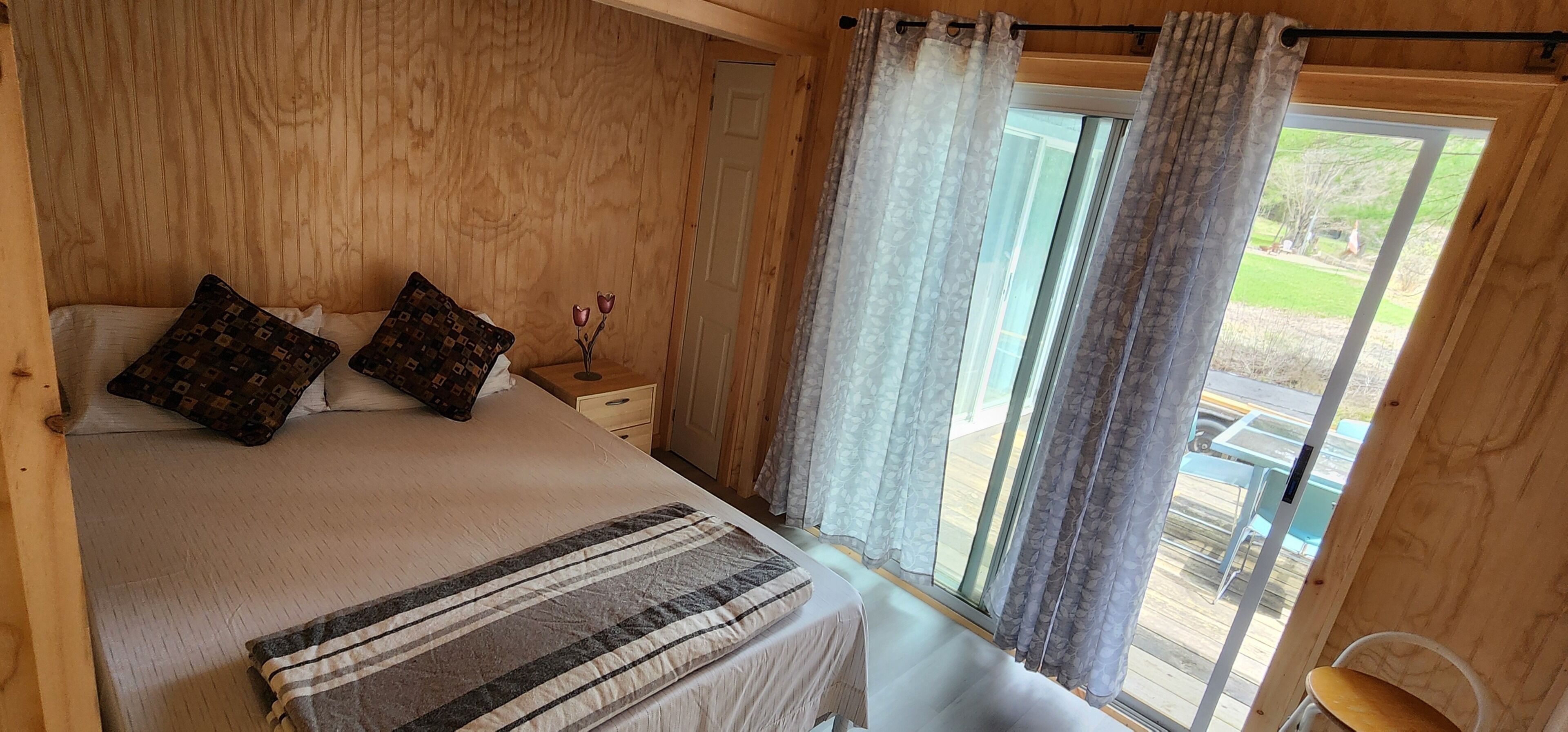 A wooden room with a bed, two pillows, a bedside table with a vase, and a window with curtains.