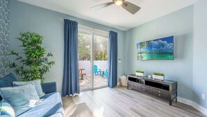 Ein Wohnzimmer mit einer Couch, einem Fernseher, einem Deckenventilator und einer Glasschiebetür, die zu einem Außenbereich mit Sitzgelegenheiten führt.