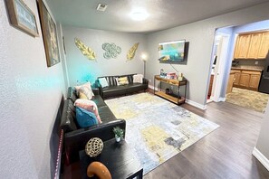 Una sala de estar con un sofá, una mesa de centro y un tapete. Hay un televisor en la pared y una cocina al fondo.