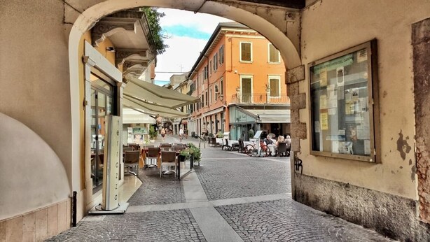 An urban street with cobblestone pavement, outdoor seating areas, and traditional buildings with balconies. - Near Gardaland, 2 minutes from the lake, in the heart of Peschiera del Garda! Xenia19 (Peschiera del Garda)