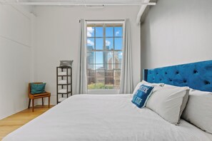 A bedroom with a large bed, a blue headboard, a window with curtains, a small wooden chair, and a metal shelf.