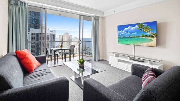 A modern living room with a large TV, grey sofas, a glass coffee table, and a balcony with a city view. - Chevron Renaissance Ocean View (Surfers Paradise)