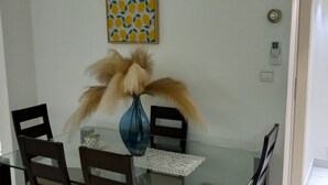 A dining area with a glass table, four chairs, a vase with dried flowers, a wall-mounted air conditioner, and a decorative wall piece.