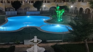 Una piscina con una fontana, circondata da un giardino curato e un edificio con archi sullo sfondo.