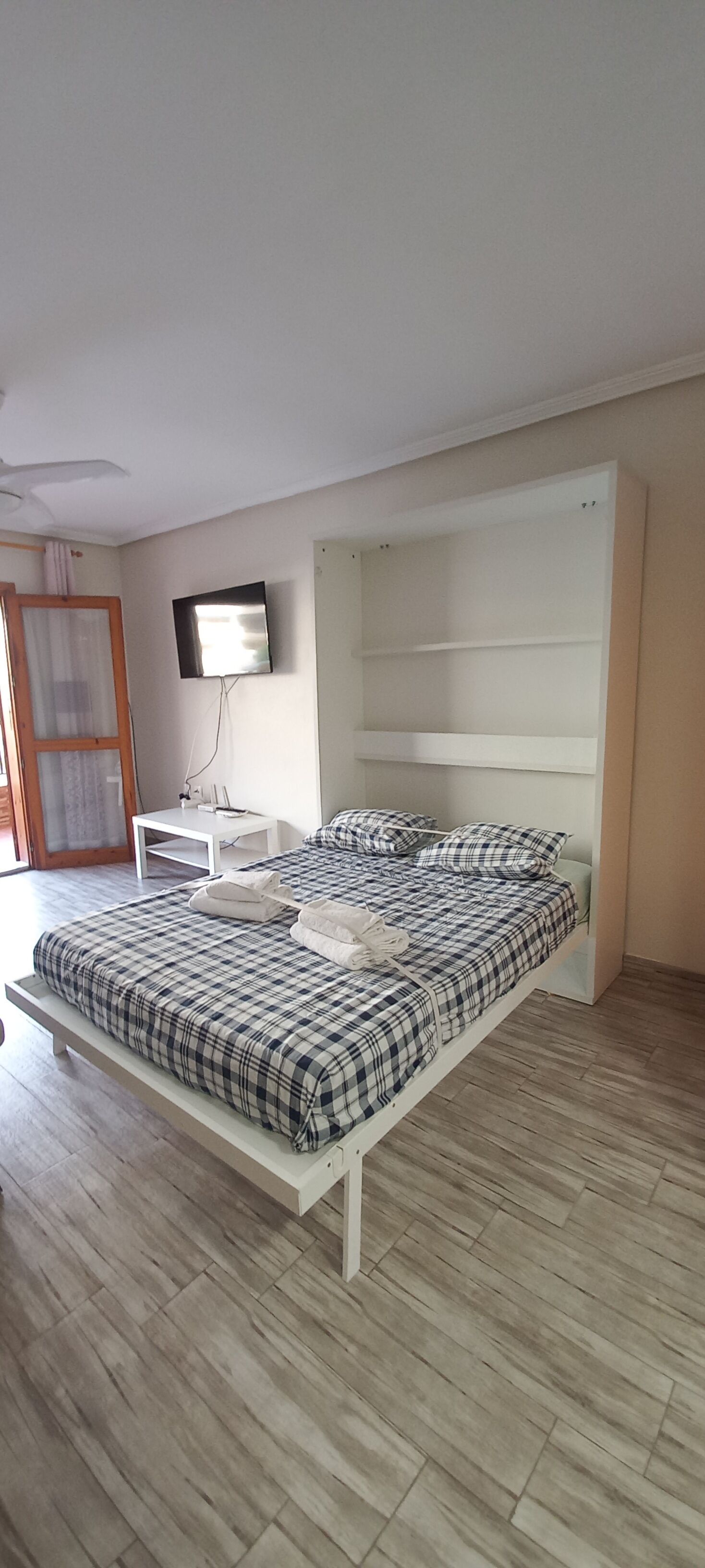 Un dormitorio con cama, televisor montado en la pared y una mesita.