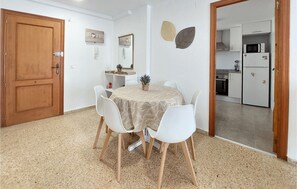 Comedor con una mesa redonda, sillas blancas y un mantel beige estampado. Se ve una puerta de madera y una cocina a través de un vano abierto.