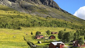 A rural landscape with a mountain backdrop, featuring several red wooden buildings, a dirt road, and scattered trees. - 3 bedroom lovely home in Hemsedal (Hemsedal)