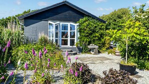 Une maison avec un toit sombre, des fenêtres à cadre blanc et un jardin agrémenté de fleurs et d’arbustes violets.