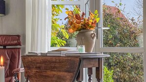 Une pièce comprenant un bureau, une chaise, un vase rempli de feuilles d’automne et une fenêtre donnant sur un paysage verdoyant et un bâtiment.