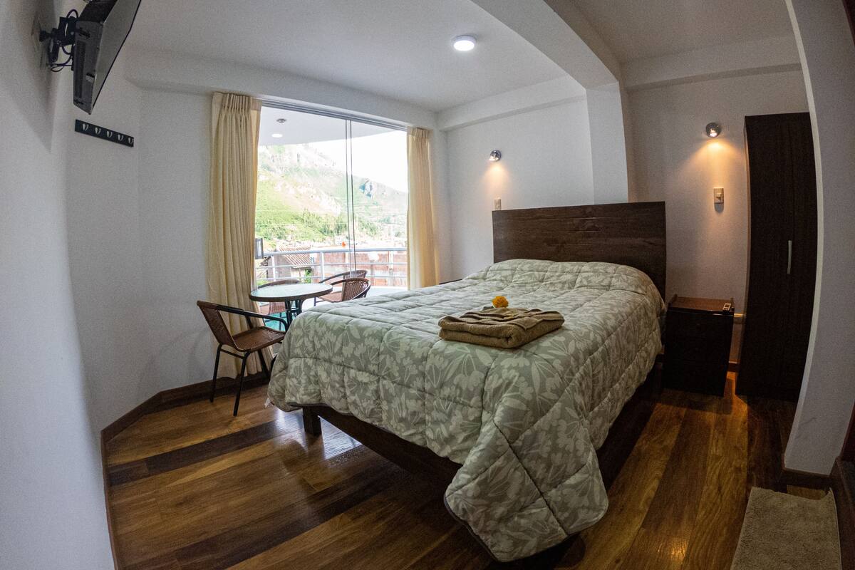 A bedroom with a bed, a small table, and chairs. There is a window with curtains, a door, and a ceiling light.