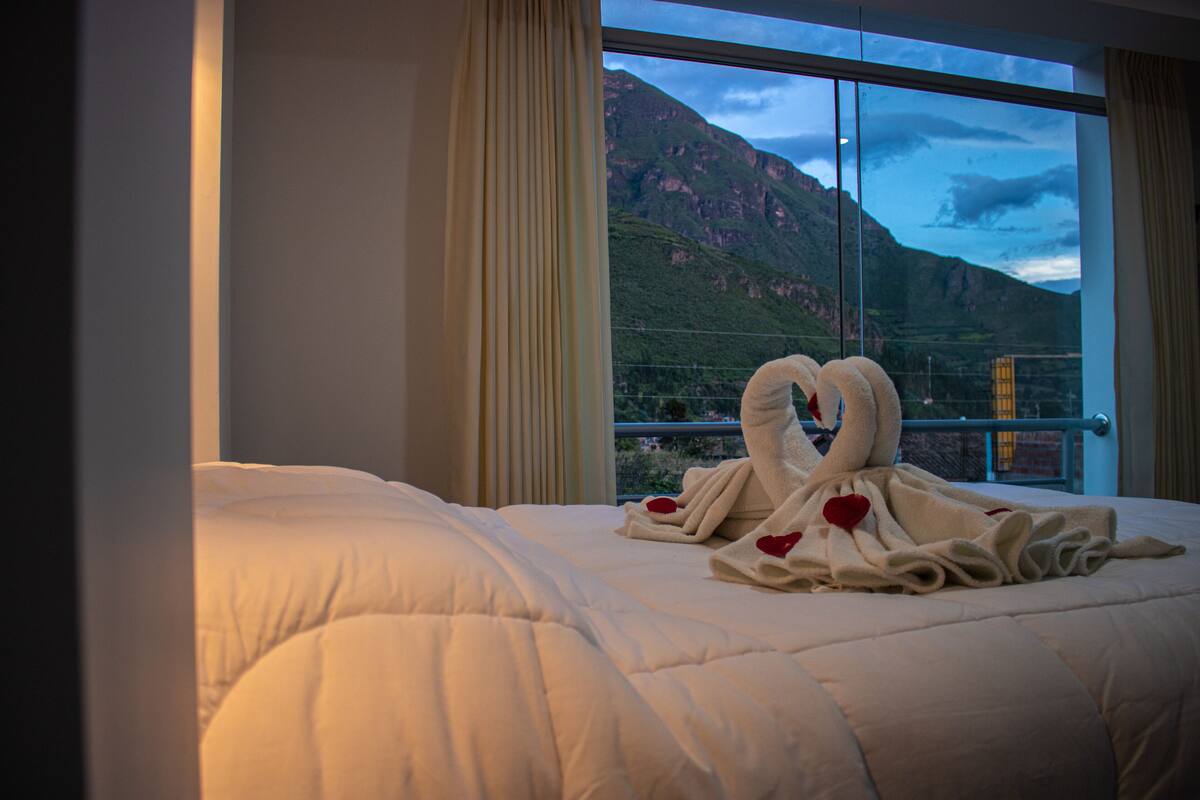 A hotel room with a large bed, a view of mountains through a window, and a towel arrangement on the bed.