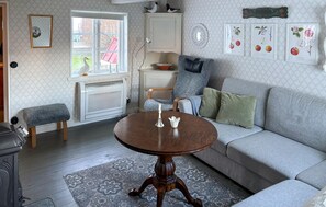 Uma sala de estar acolhedora com um sofá cinzento, uma mesa de centro de madeira e um tapete estampado. Há também um radiador branco, um banco e uma janela com vista para o exterior.