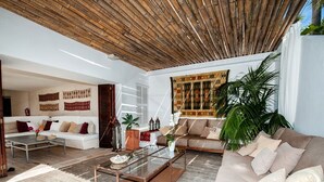 Uma sala de estar moderna com teto de madeira, um sofá branco, uma mesa de centro de vidro e uma grande planta em vaso.