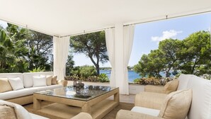 Uma área de estar moderna ao ar livre, com mesa de centro de vidro, sofás bege e vista para o mar.