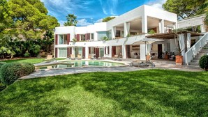 Uma casa moderna de dois andares com piscina, cercada por vegetação exuberante e um céu azul límpido.