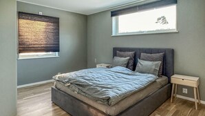 A bedroom with a bed, a nightstand, a window with blinds, and a view of trees outside.