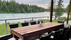 Un área de comedor al aire libre con mesa de madera y sillas de mimbre, con vista a un río y un área boscosa.