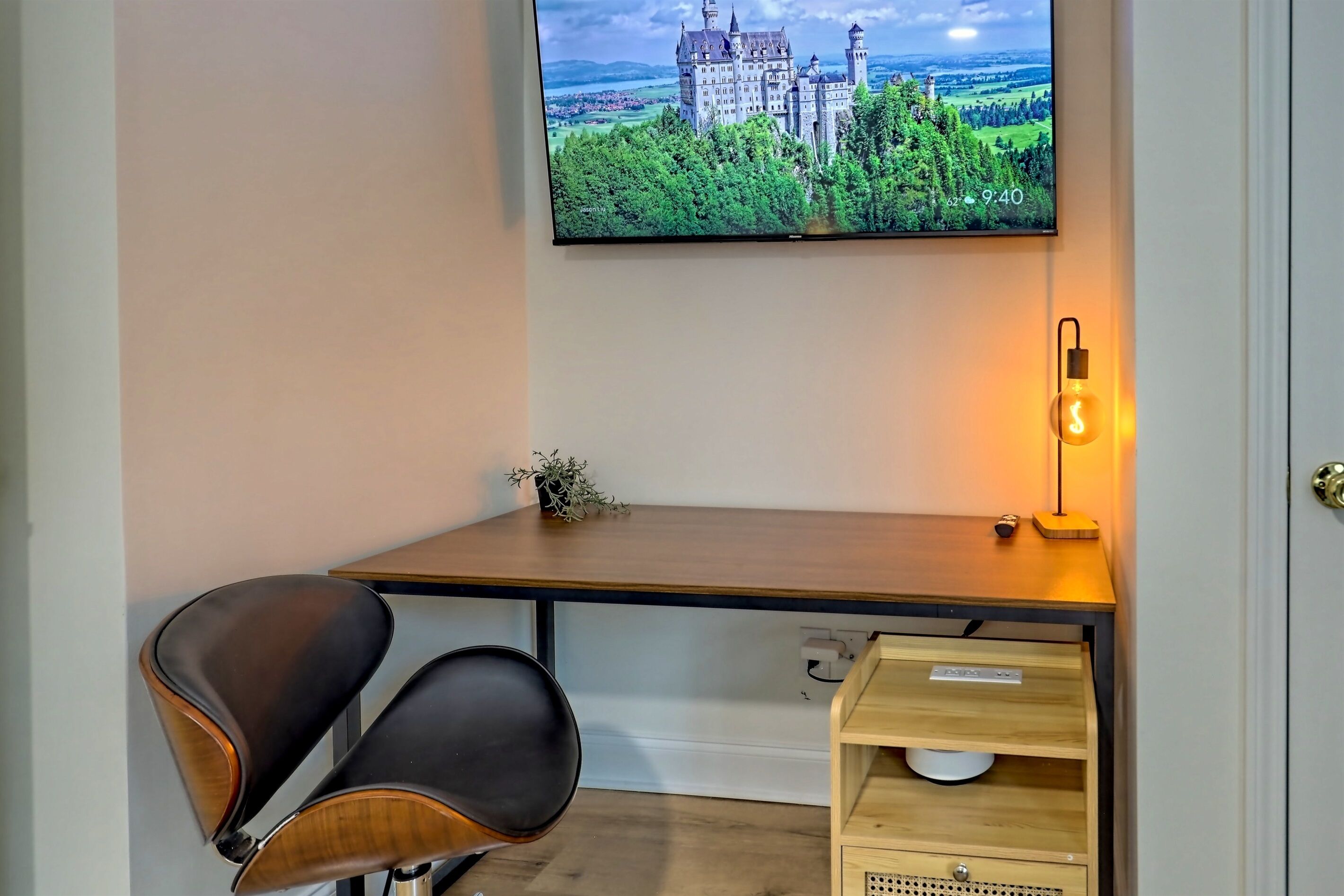 A modern room with a wooden desk, two black chairs, a television displaying a castle, and a bedside table with a lamp.