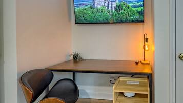 A modern room with a wooden desk, two black chairs, a television displaying a castle, and a bedside table with a lamp.