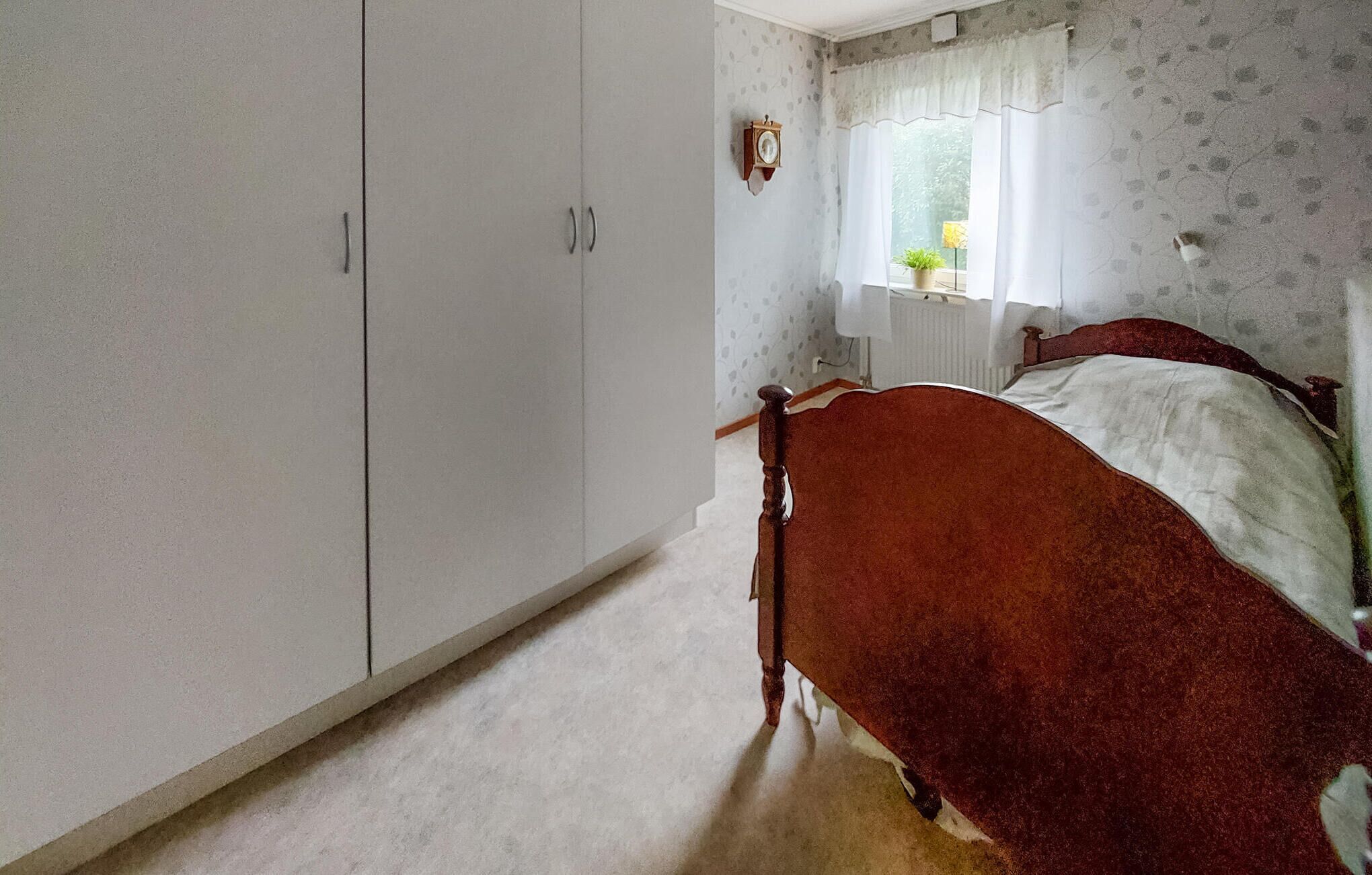 A bedroom with a large wardrobe, a bed with a wooden headboard, a window with curtains, and a wall clock.