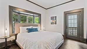 A bedroom with a large bed, a window, a door, and a small table with a lamp.