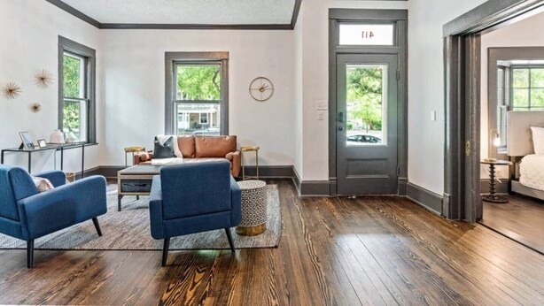 A spacious living room with a large window, a brown leather sofa, a blue armchair, and a wooden floor.