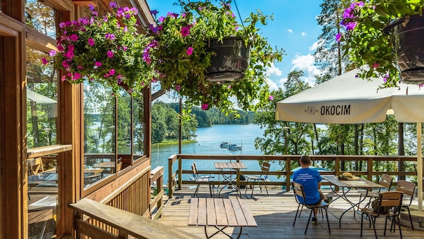 Terraza de madera con mesas y sillas con vista a un lago, adornada con cestas de flores colgantes y un área cubierta con una sombrilla que dice "OKOCIM".