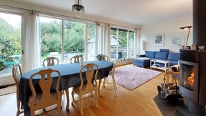 Ein großzügiges Wohnzimmer mit Essbereich, großen Fenstern, einem Kamin und Blick ins Freie.