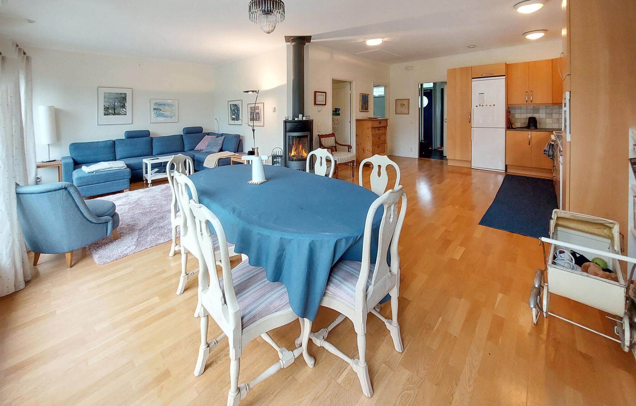 En rummelig stue med blå sofaarrangement, et spisebord med hvide stole, en pejs og et køkkenområde med træskabe.