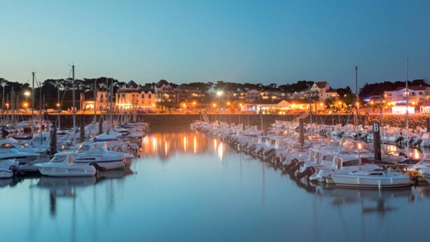 Uma marina ao entardecer, com barcos atracados e edifícios ao fundo.