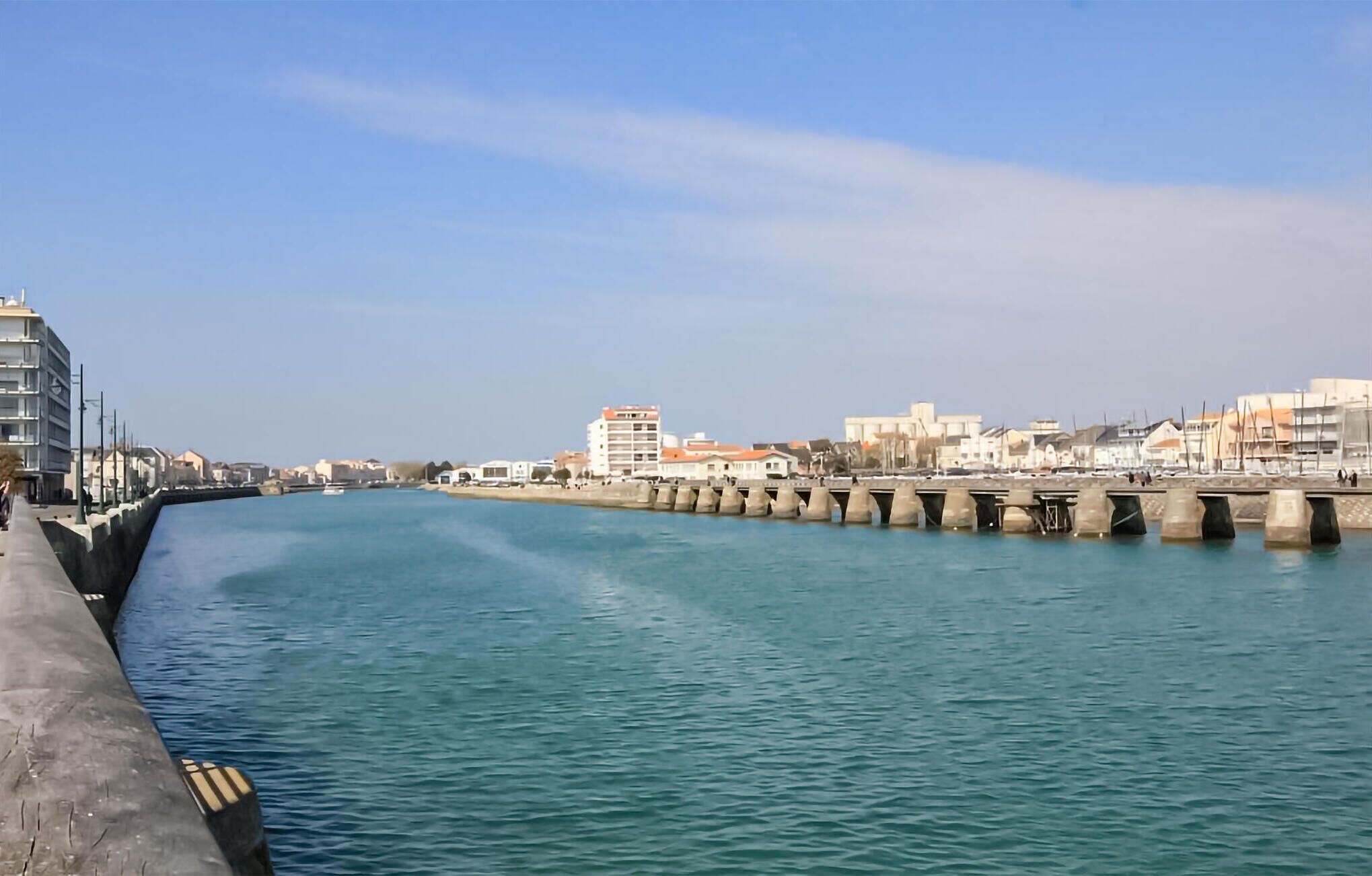 Une promenade en bord de mer, avec une rivière paisible, une barrière en béton et des bâtiments en arrière-plan.