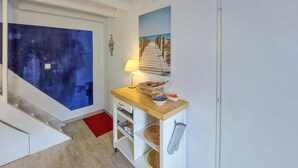 Um corredor com escada de madeira, um quadro abstrato azul, uma mesa lateral de madeira com abajur e uma fotografia emoldurada de um calçadão de praia.