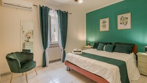 A bedroom with a bed, a chair, a nightstand, a lamp, and two framed botanical prints on the wall.