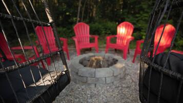 Una silla colgante de mimbre, un pozo de fuego y sillas Adirondack rojas en un jardín.