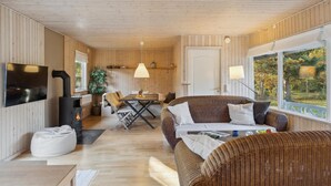 A cozy cabin interior with a dining area, a fireplace, wicker furniture, and a large window offering a view of the outdoors. - Amazing home in Nykøbing Sj with WiFi (Nykøbing Sj)
