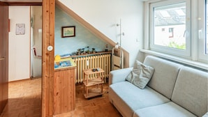 Une pièce chaleureuse avec un plancher en bois, un canapé gris, un banc en bois, un radiateur et une fenêtre donnant sur une maison.