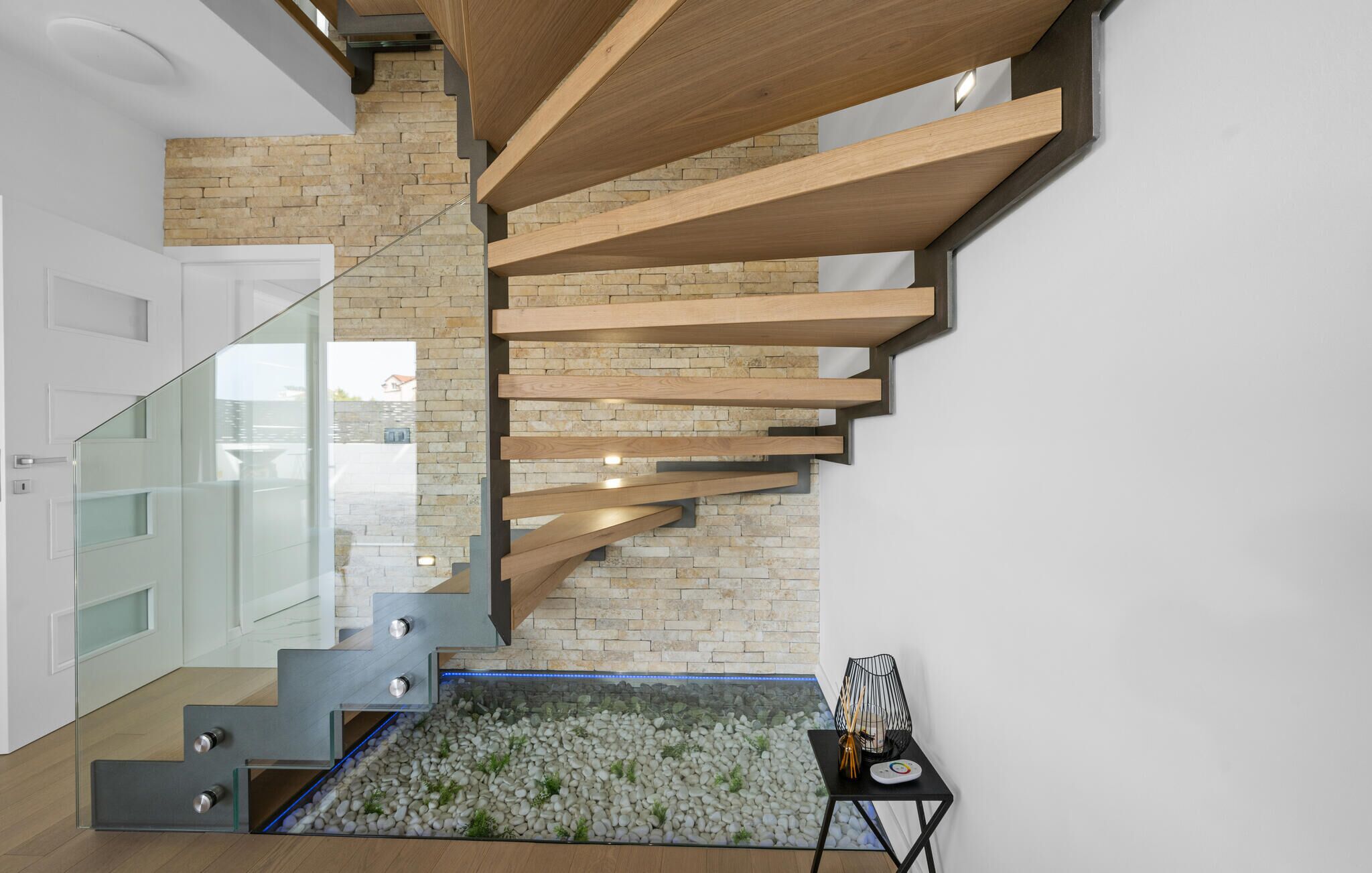 Een moderne trap met houten balken en een glazen balustrade, die leidt naar een kamer met een klein tafeltje en decoratieve objecten.