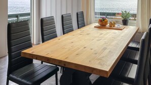 Une table à manger entourée de six chaises noires, avec un plateau en bois, et une vue sur la mer grâce à de grandes fenêtres.