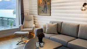 Un salon chaleureux avec un canapé, un fauteuil et une table agrémentée d’un vase et de bougeoirs. Un tableau est accroché au mur, et une fenêtre offre une vue sur un chalet et des montagnes.