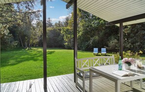 En overdækket terrasse med træterrasse, et hvidt bord med pude og to stribede stole.