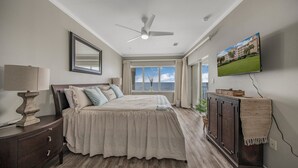 Une chambre à coucher comprenant un lit, une table de chevet, une commode, un ventilateur de plafond, un téléviseur et une fenêtre donnant sur l’océan.
