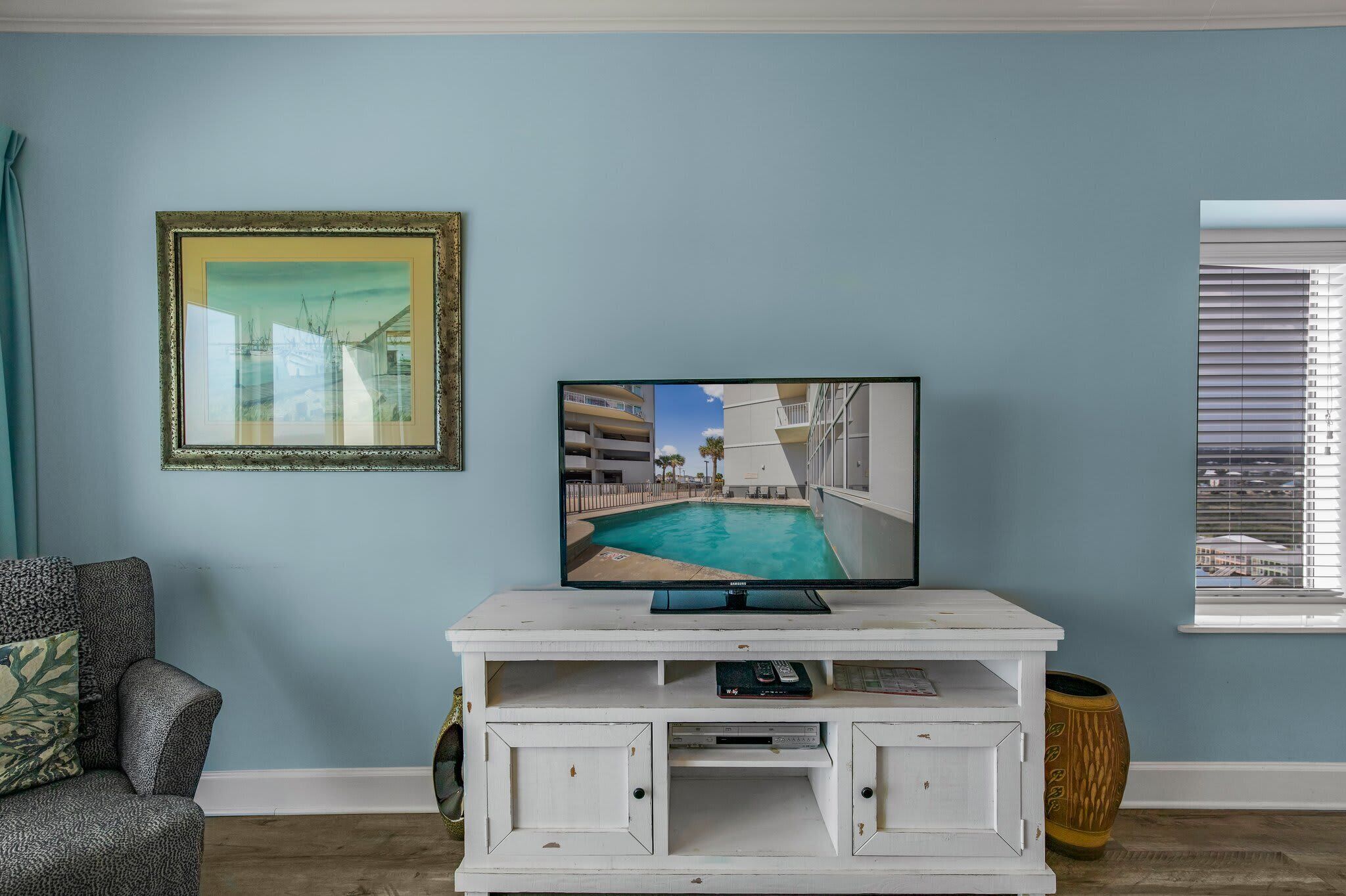 A living room with a TV on a white entertainment unit, a framed picture on the wall, and a window with blinds.