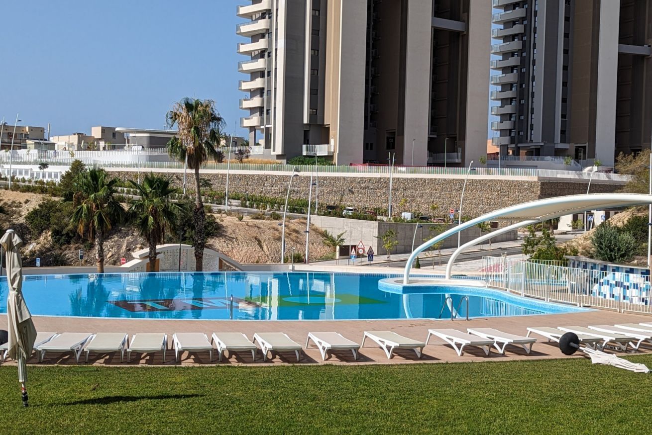 En swimmingpool med liggestole, palmer og moderne bygninger i baggrunden.
