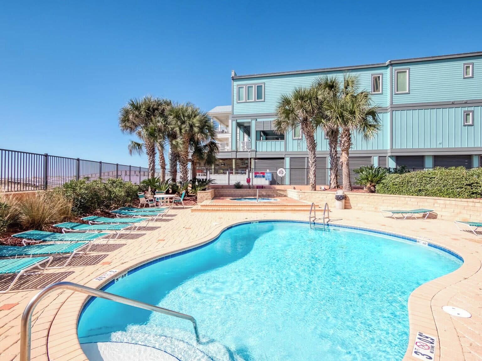 A swimming pool with lounge chairs and palm trees, surrounded by a building with a light blue exterior.