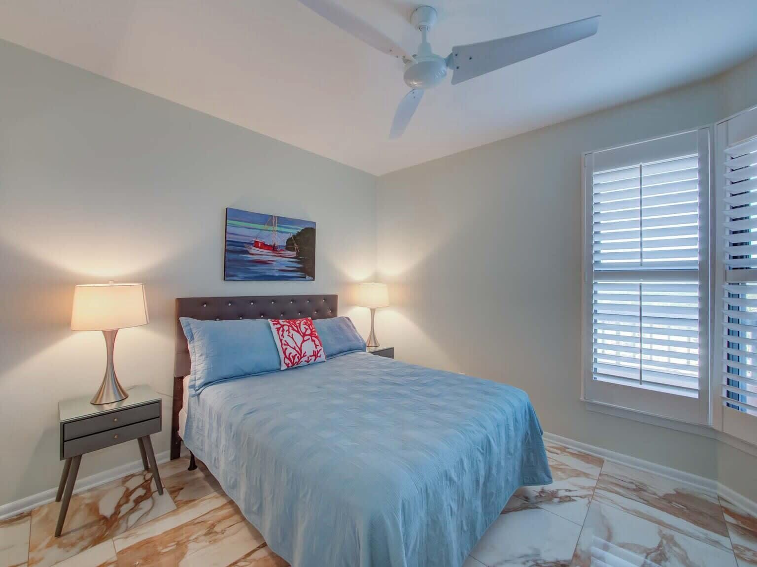 A bedroom with a bed, two bedside lamps, a ceiling fan, a painting, and a window with blinds.