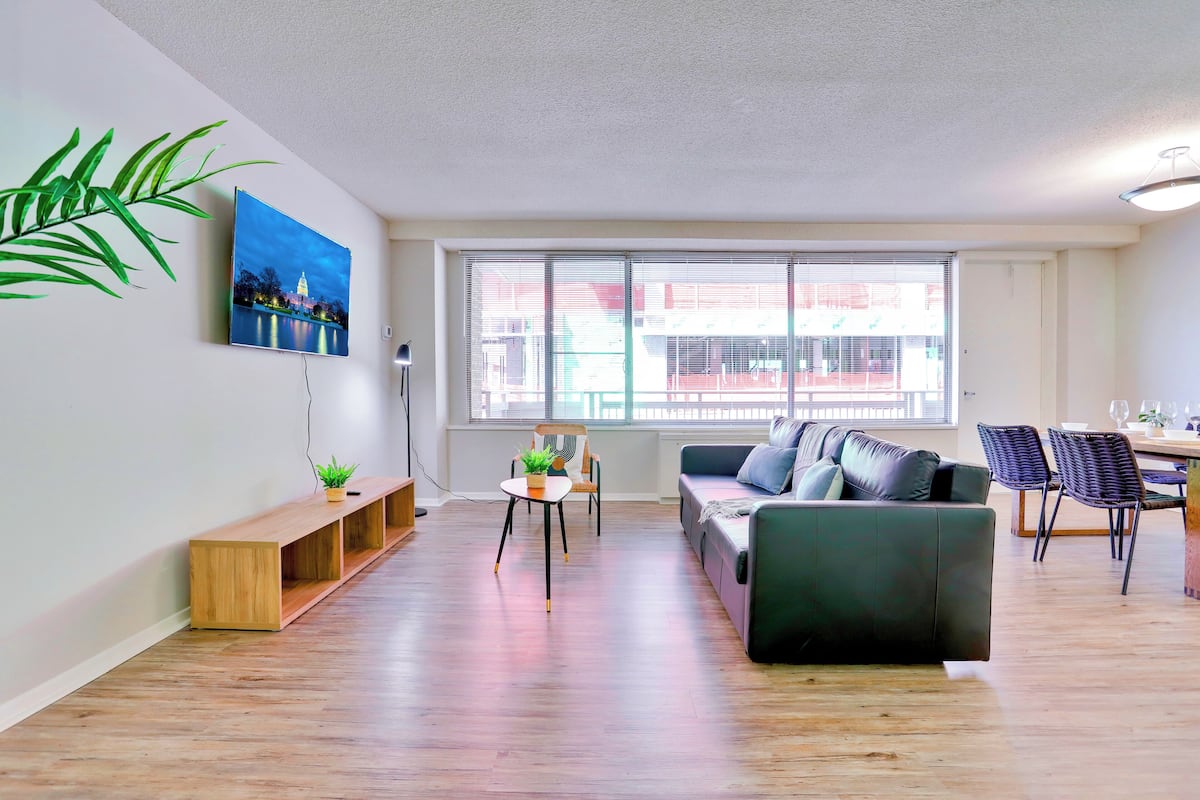 Een moderne woonkamer met een bank, een eettafel en stoelen. Een groot raam biedt uitzicht op gebouwen.