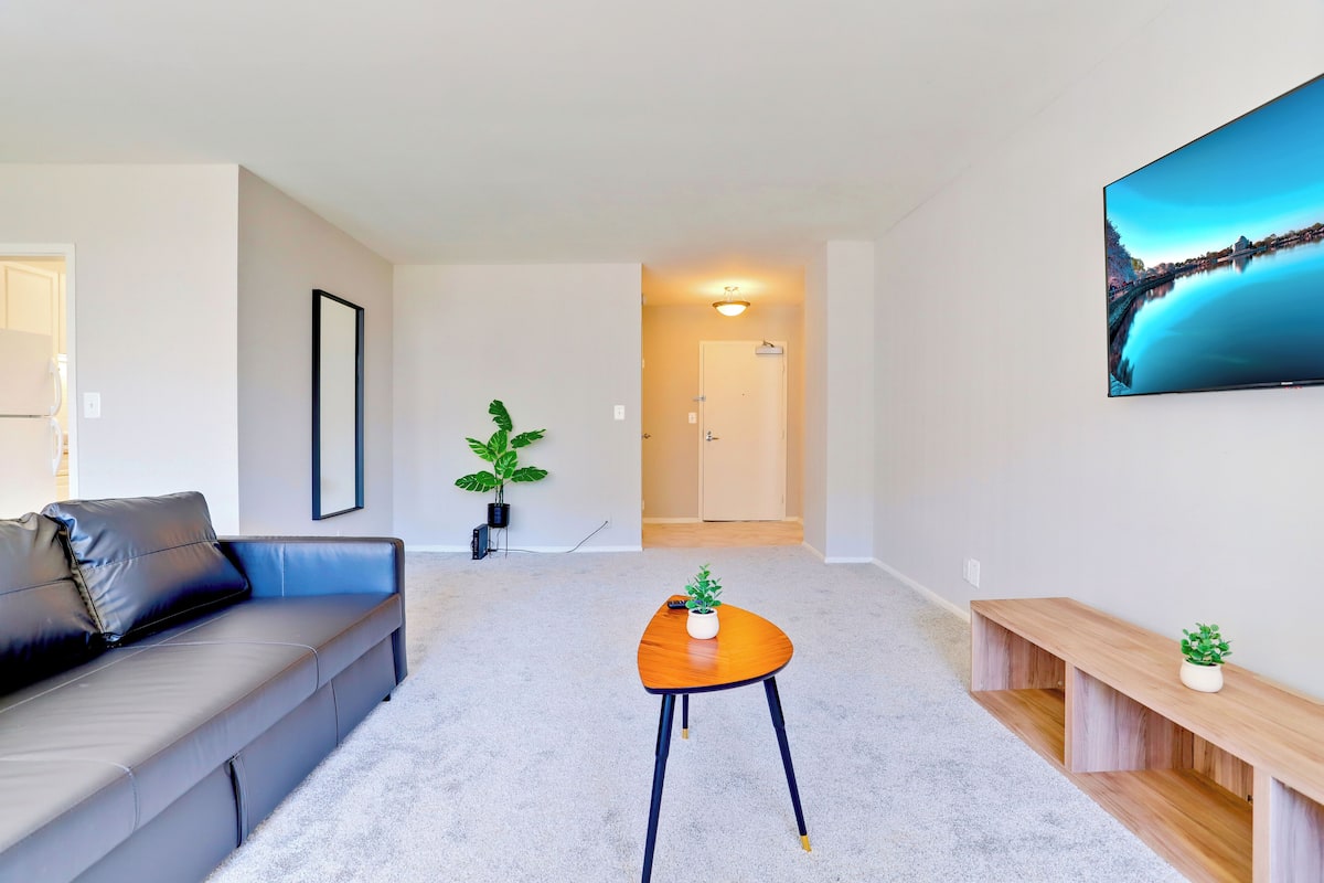 Een moderne woonkamer met een grijze bank, een houten salontafel en een houten bankje. Op de salontafel staat een potplant en op het bankje ook. Een ingelijst schilderij hangt aan de muur, en aan de muur is een flatscreen televisie bevestigd.